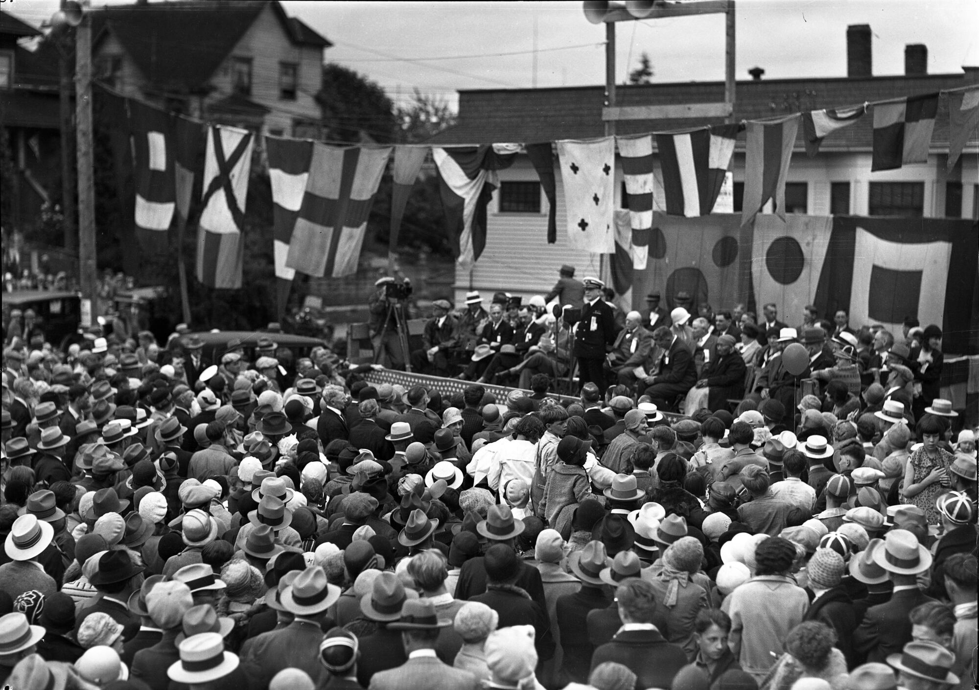 A large crowd in hats gathers at the Manette Bridge dedication in Bremerton in 1930 as a uniformed officer speaks from a flag-draped stand