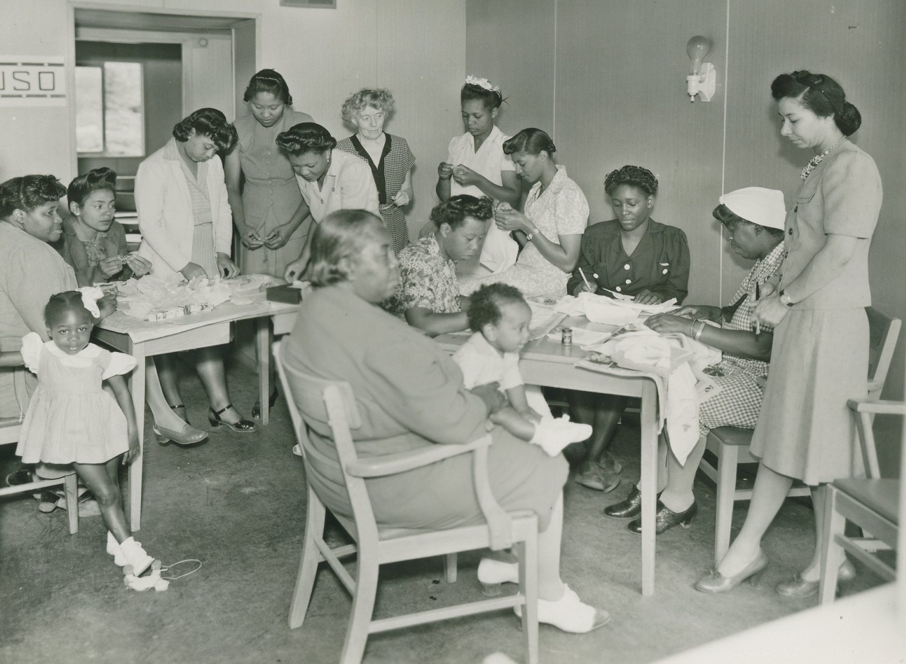 A group of Black women sew at tables in a USO room while two small children look on
