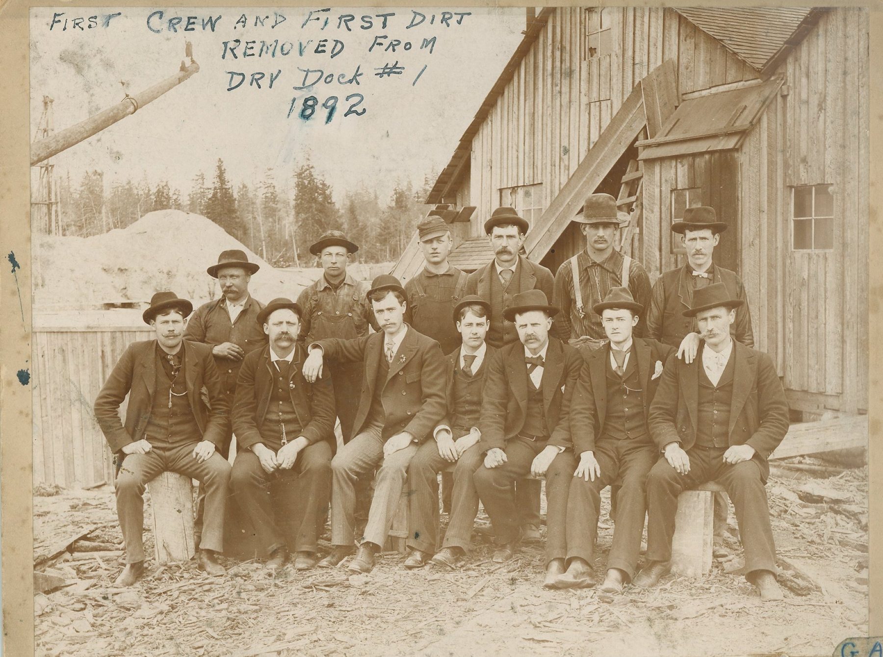 About a dozen men in hats and work clothes pose on logs in front of a plank building