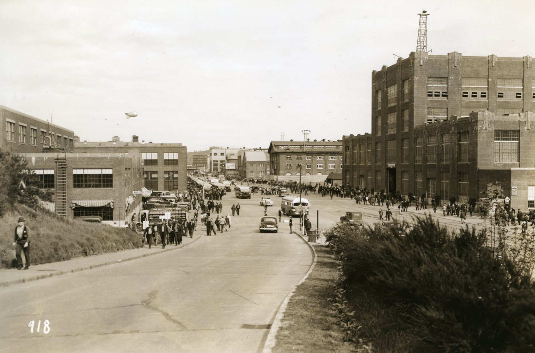 Crowds of workers, buses and cars at the Navy Yard entrance between brick buildings, with a barrage balloon in the sky