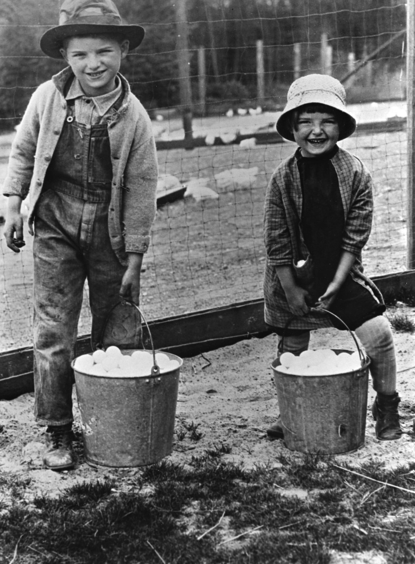 Two children in work clothes stand holding buckets full of eggs