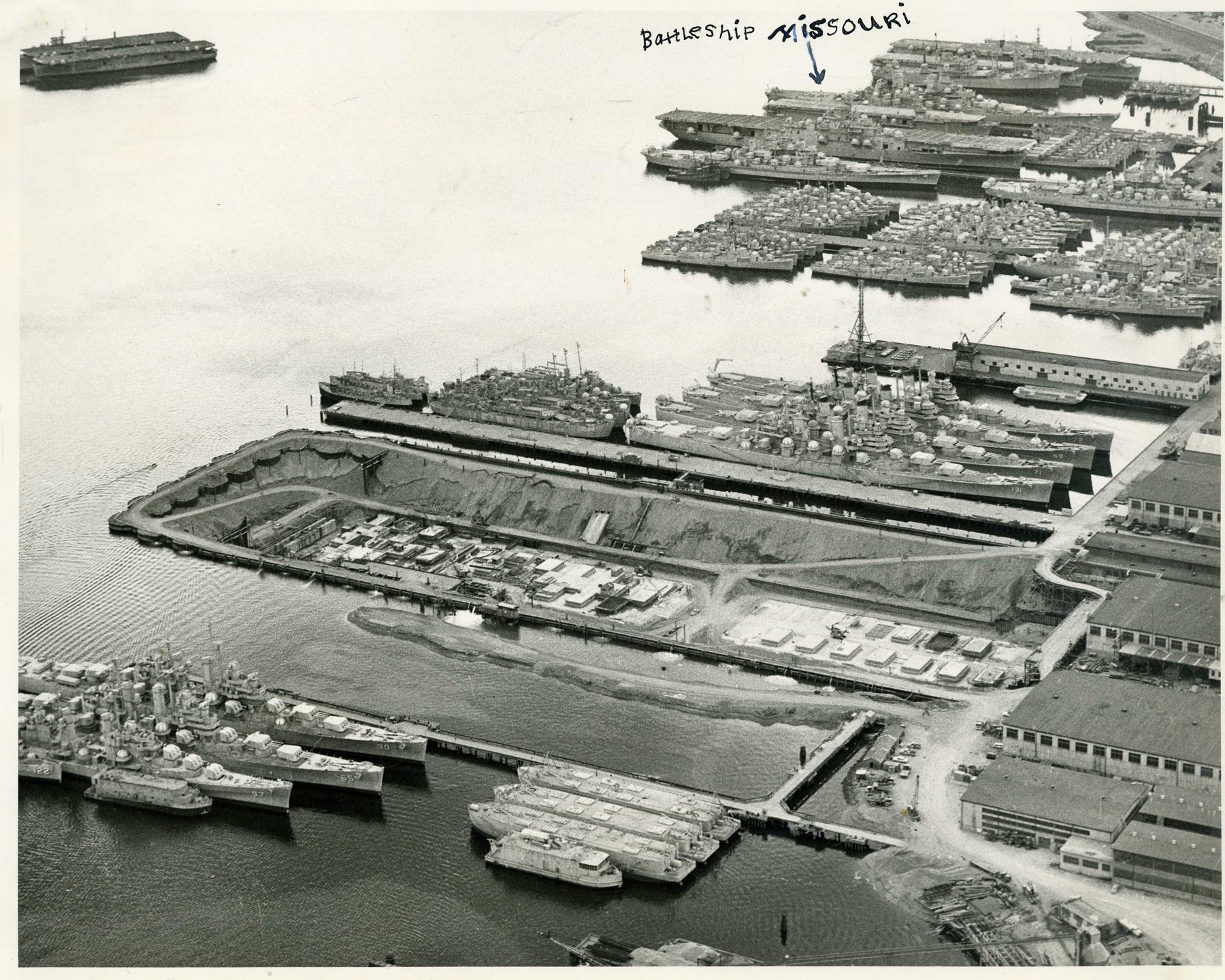 Aerial view of a shipyard dry dock with rows of gray reserve-fleet ships moored alongside