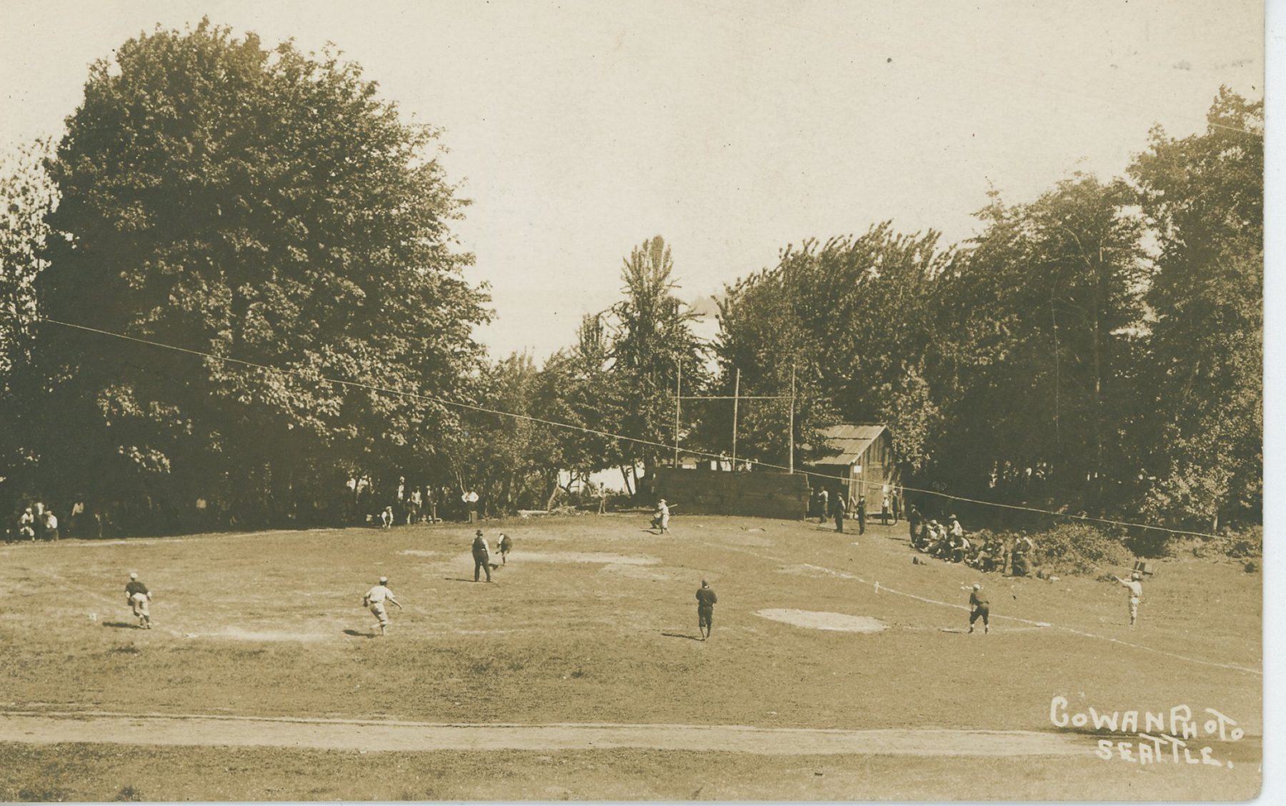 Players and spectators at a baseball game on a grassy field ringed by tall trees