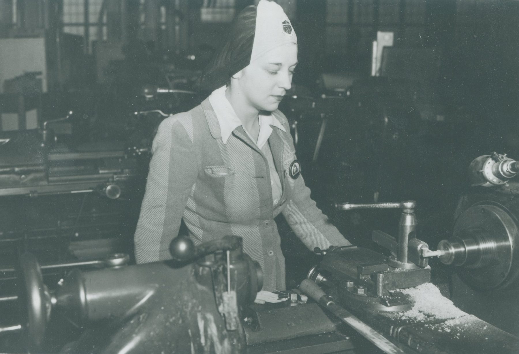A woman in a Navy Yard cap works at a lathe in a machine shop